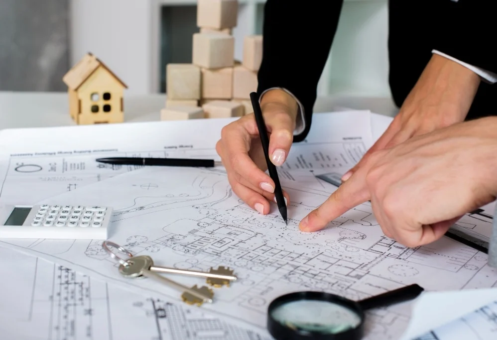 A close-up of professionals pointing to architectural blueprints on a desk with a calculator, house keys, and a magnifying glass to prepare a detailed Bill of Quantities for a construction project.