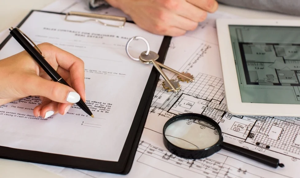 A person signing a formal construction contract or Bill of Quantity document next to a digital tablet displaying floor plans, used to finalize materials and costs in a building project.